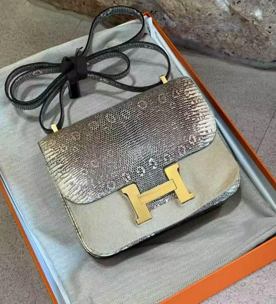 A luxury handbag with a snakeskin-patterned flap and a solid beige body, featuring a prominent gold H-shaped clasp and a black shoulder strap, resting inside an orange and beige box.
