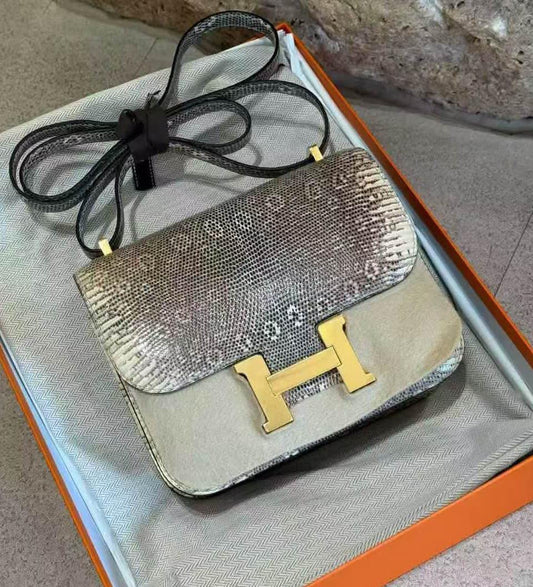 A luxury handbag with a snakeskin-patterned flap and a solid beige body, featuring a prominent gold H-shaped clasp and a black shoulder strap, resting inside an orange and beige box.